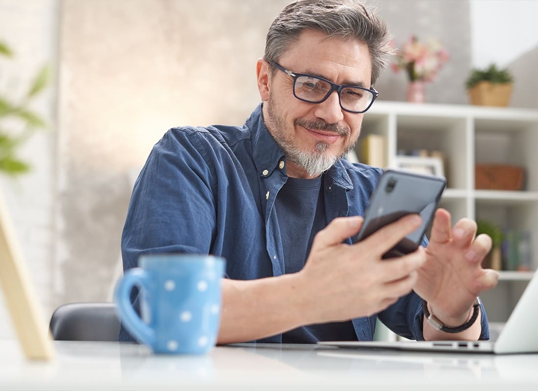 Read Our Reviews - Middle Aged Man Scrolling Through His Smartphone While Smiling With His Blue Coffee Cup Next to His Laptop